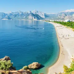 Słoneczna plaża na Riwierze Tureckiej, z turkusowym morzem, górami w tle i ludźmi odpoczywającymi na piasku.