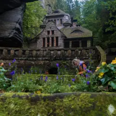 Kamienny domek w otoczeniu skał i zieleni, idealne miejsce na czeska szwajcaria noclegi. Widać kwitnące kwiaty i osobę pochylającą się nad nimi.