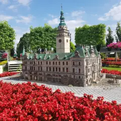 Miniatura ratusza z wieżą zegarową, otoczona czerwonymi kwiatami, przypomina o architekturze Kotliny Kłodzkiej.