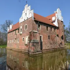 Zamek otoczony fosą, z ceglanymi murami i charakterystycznymi białymi szczytami. Woda odbija jego architekturę, tworząc malowniczy widok. Miękinia.
