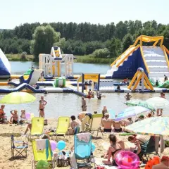 Rodzinny wypoczynek nad wodą na **bolmin zalew** z dmuchanymi atrakcjami wodnymi, zjeżdżalniami i placem zabaw. Słoneczny dzień, plaża pełna ludzi.