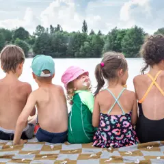 Szczęśliwe wakacje nad jeziorem z dziećmi. Grupa dzieci w strojach kąpielowych siedzi na kocu, ciesząc się słońcem i widokiem na wodę.