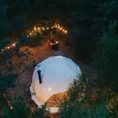 Nocny relaks w lesie. Biała kopuła glampingu w Kujawsko-Pomorskiem, oświetlona girlandami, z paleniskiem w tle.