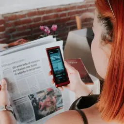 Kobieta z rudymi włosami używa telefonu do tłumaczenia tekstu z gazety. To przydatna wskazówka, jak poradzić sobie na lotnisku bez znajomości języka.