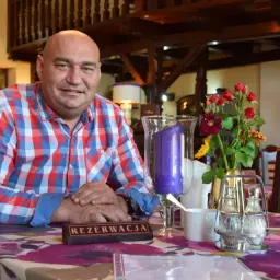 Łysy mężczyzna w kratę siedzi przy stole w karczmie. Na stole wazon z kwiatami i świeca.