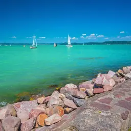 Balaton wczasy z wyżywieniem: żaglówki na turkusowej wodzie, kamienisty brzeg i błękitne niebo. Idealne miejsce na relaks.