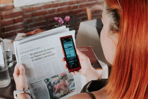 Kobieta z rudymi włosami używa telefonu do tłumaczenia tekstu z gazety. To przydatna wskazówka, jak poradzić sobie na lotnisku bez znajomości języka.