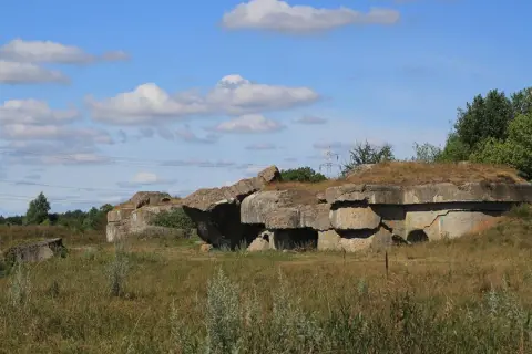 Ruiny fortu Osowiec, betonowe bunkry porośnięte trawą, na tle błękitnego nieba z chmurami.