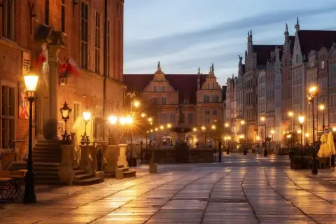 Długa ulica w Gdańsku oświetlona latarniami, z zabytkowymi kamienicami i fontanną.