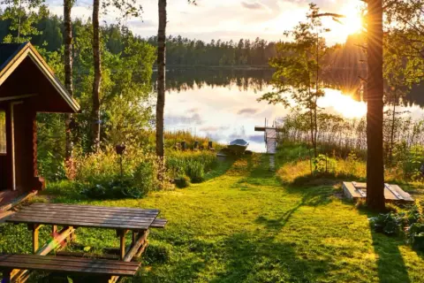 Letnia chata nad jeziorem w Finlandii. Słońce zachodzi, rzucając złote światło na trawę i wodę. Ceny w Finlandii mogą być wysokie, ale widoki są bezcenne.
