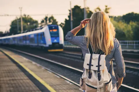 Dziewczyna z plecakiem czeka na pociąg nad morze. Słońce oświetla jej blond włosy.