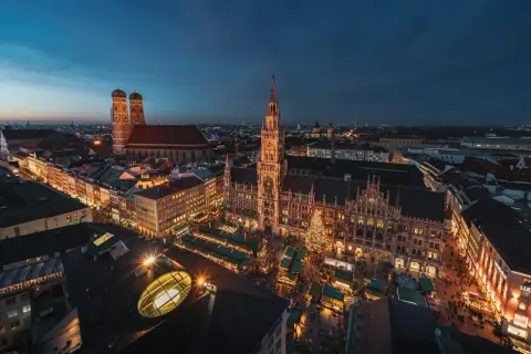 Nocny jarmark bożonarodzeniowy w Monachium. Rynek z oświetlonymi straganami i choinką, w tle majestatyczny ratusz i katedra.