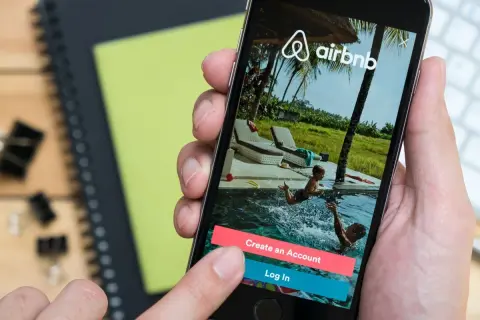 Dłoń trzyma smartfon z aplikacją Airbnb. Widok na basen z dziećmi. Zastanawiasz się airbnb co to? To platforma do rezerwacji noclegów.