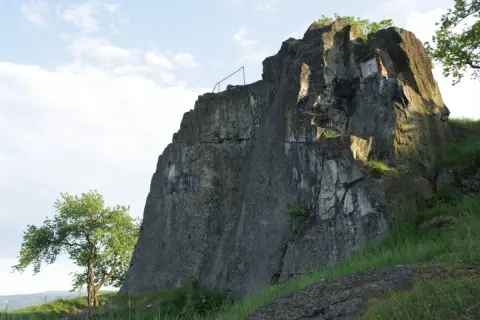Skaliste zbocze z widocznymi szczelinami i fragmentem muru, porośnięte trawą. Na tle błękitnego nieba z chmurami. W oddali drzewo.