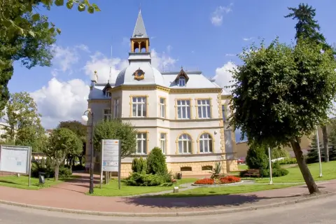 Elegancki budynek w Dębnie z wieżyczką i ozdobnymi detalami, otoczony zielenią i kwitnącymi rabatami.