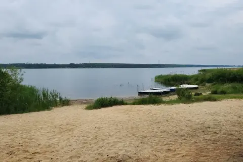 Piękny, piaszczysty brzeg jeziora w Poraju. Kilka łódek czeka na wodzie, otoczone zielenią.