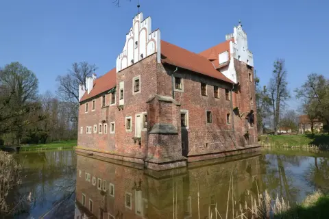 Zamek otoczony fosą, z ceglanymi murami i charakterystycznymi białymi szczytami. Woda odbija jego architekturę, tworząc malowniczy widok. Miękinia.