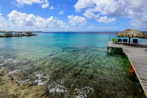 Bonaire wyspa: drewniany pomost z leżakami i parasolem, turkusowa woda, skaliste dno, chmury na niebie.