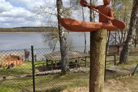 Drewniana rzeźba kajakarza nad jeziorem Szczytno. W tle widać taflę wody, las i zabudowania.