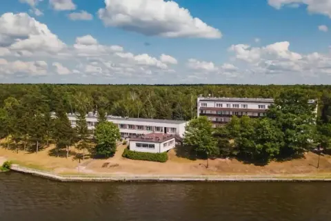 Budynki hotelowe w Ryni nad jeziorem, otoczone lasem i zielenią.