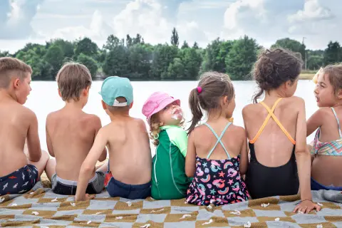 Szczęśliwe wakacje nad jeziorem z dziećmi. Grupa dzieci w strojach kąpielowych siedzi na kocu, ciesząc się słońcem i widokiem na wodę.