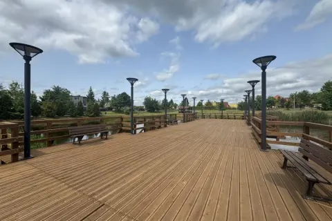 Drewniana promenada w Swarożynie z ławkami i latarniami, nad spokojną wodą, pod błękitnym niebem z chmurami.