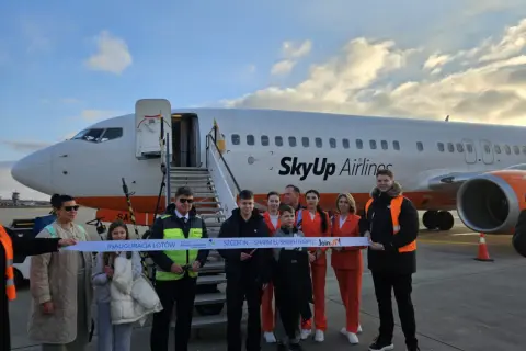 Grupa osób przecina wstęgę przed samolotem SkyUp Airlines. Inauguracja lotów do Szarm El-Szejk. Lotnisko nad morzem.