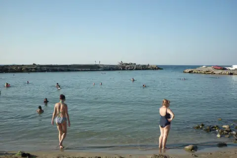 Dwie kobiety w strojach kąpielowych stoją na brzegu morza. W tle widać kamienne **gouves** i ludzi kąpiących się w wodzie.