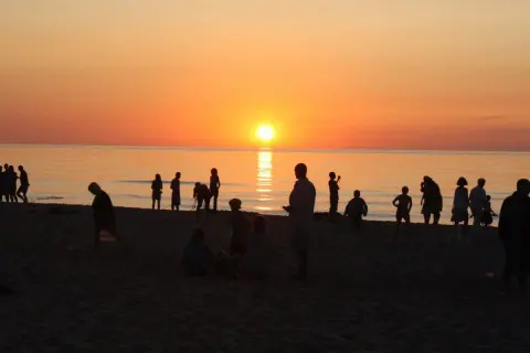 Ludzie na plaży podziwiają piękny zachód słońca w Sarbinowie. Słońce odbija się w spokojnym morzu, tworząc złocistą poświatę.