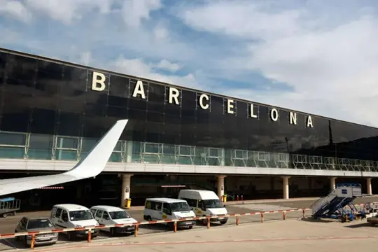 Duży napis BARCELONA nad nowoczesnym terminalem lotniska. Samolotowy skrzydło i busy czekają na pasażerów.