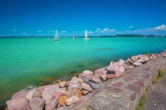 Balaton wczasy z wyżywieniem: żaglówki na turkusowej wodzie, kamienisty brzeg i błękitne niebo. Idealne miejsce na relaks.