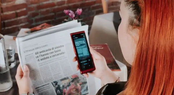 Kobieta z rudymi włosami używa telefonu do tłumaczenia tekstu z gazety. To przydatna wskazówka, jak poradzić sobie na lotnisku bez znajomości języka.