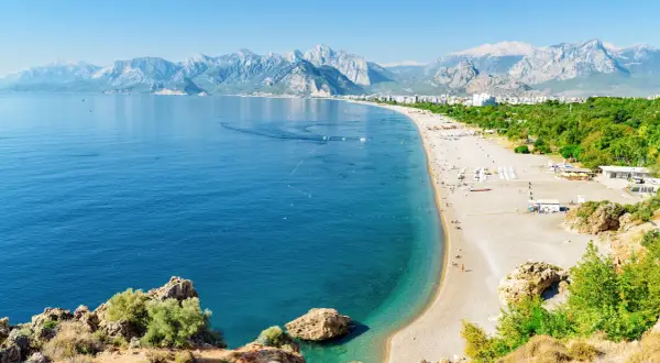 Słoneczna plaża na Riwierze Tureckiej, z turkusowym morzem, górami w tle i ludźmi odpoczywającymi na piasku.
