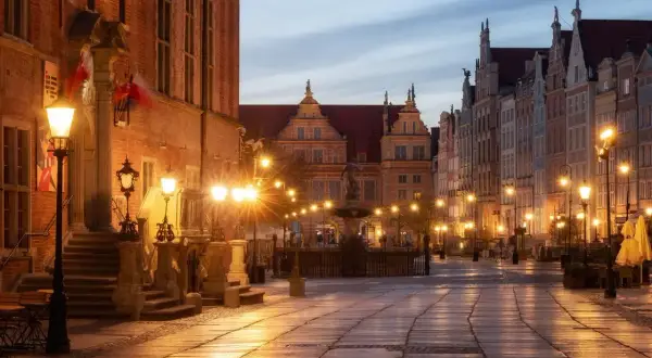 Długa ulica w Gdańsku oświetlona latarniami, z zabytkowymi kamienicami i fontanną.