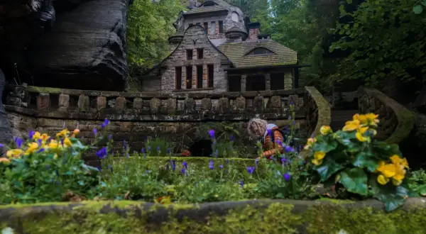 Kamienny domek w otoczeniu skał i zieleni, idealne miejsce na czeska szwajcaria noclegi. Widać kwitnące kwiaty i osobę pochylającą się nad nimi.