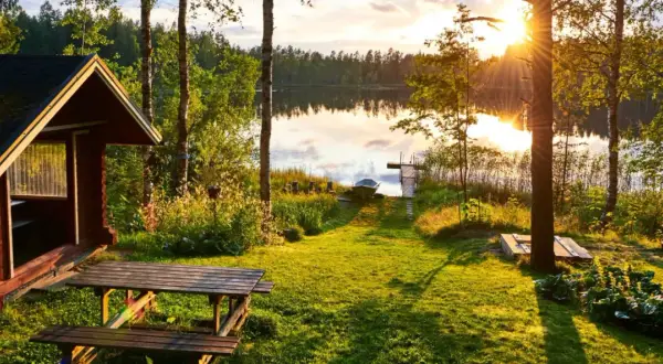 Letnia chata nad jeziorem w Finlandii. Słońce zachodzi, rzucając złote światło na trawę i wodę. Ceny w Finlandii mogą być wysokie, ale widoki są bezcenne.