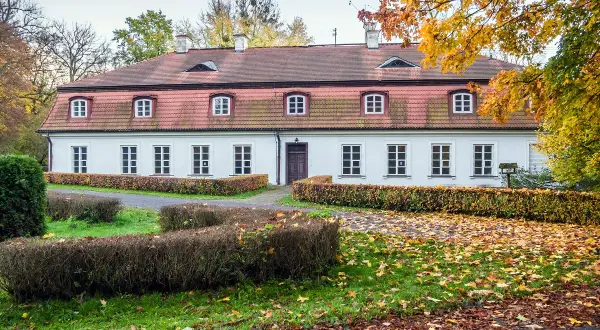 Biały dworek z czerwonym dachem, otoczony jesiennymi drzewami i krzewami. Wokół leżą opadłe liście, tworząc malowniczy krajobraz. Dawniej w takich miejscach znajdowały się obory.