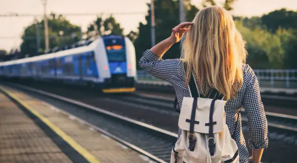 Dziewczyna z plecakiem czeka na pociąg nad morze. Słońce oświetla jej blond włosy.