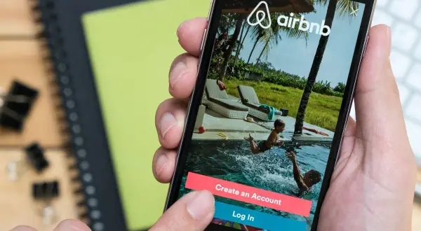 Dłoń trzyma smartfon z aplikacją Airbnb. Widok na basen z dziećmi. Zastanawiasz się airbnb co to? To platforma do rezerwacji noclegów.
