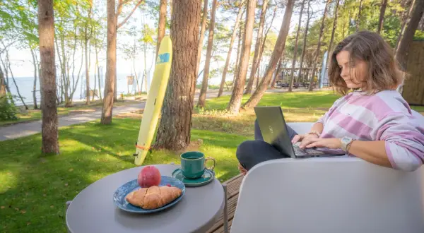 Kobieta pracuje na laptopie przy stoliku z kawą i croissantem. W tle widać morze i sosny, idealne miejsce dla domków z wyjściem na plażę.