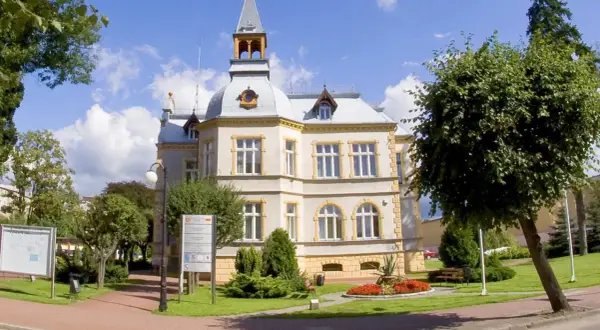 Elegancki budynek w Dębnie z wieżyczką i ozdobnymi detalami, otoczony zielenią i kwitnącymi rabatami.