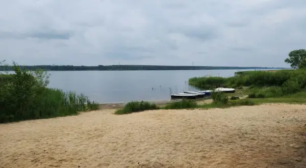 Piękny, piaszczysty brzeg jeziora w Poraju. Kilka łódek czeka na wodzie, otoczone zielenią.