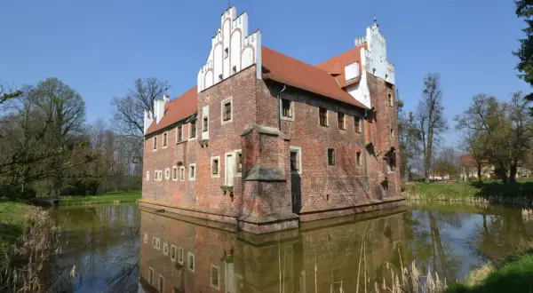 Zamek otoczony fosą, z ceglanymi murami i charakterystycznymi białymi szczytami. Woda odbija jego architekturę, tworząc malowniczy widok. Miękinia.