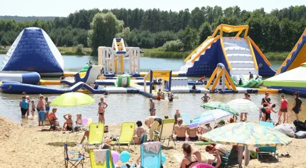 Rodzinny wypoczynek nad wodą na **bolmin zalew** z dmuchanymi atrakcjami wodnymi, zjeżdżalniami i placem zabaw. Słoneczny dzień, plaża pełna ludzi.