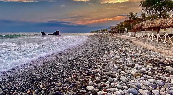 Kamienista plaża w Salvadorze o zachodzie słońca. Fale obmywają brzeg, a w oddali widać skały.