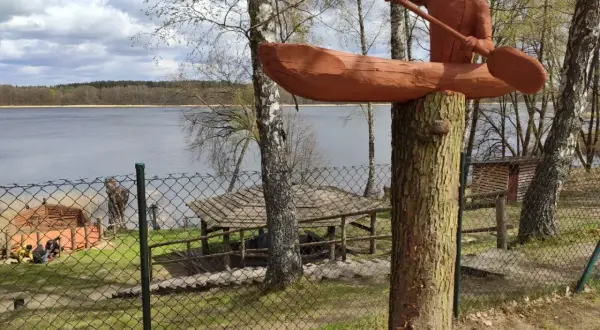 Drewniana rzeźba kajakarza nad jeziorem Szczytno. W tle widać taflę wody, las i zabudowania.