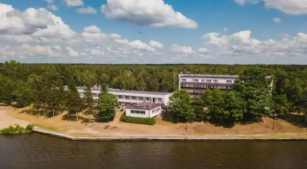Budynki hotelowe w Ryni nad jeziorem, otoczone lasem i zielenią.