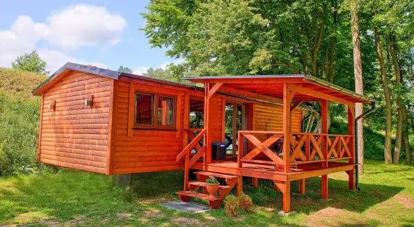 Tanie domki letniskowe nad jeziorem, drewniane, z werandą i schodkami, otoczone zielenią i drzewami.