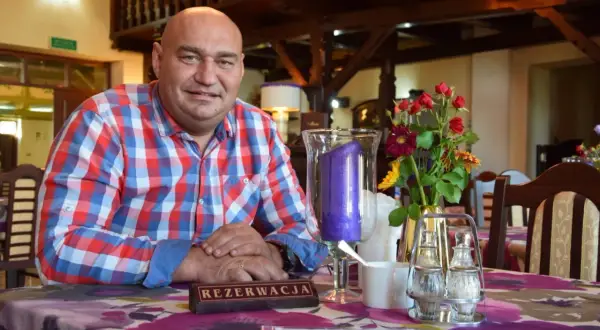 Łysy mężczyzna w kratę siedzi przy stole w karczmie. Na stole wazon z kwiatami i świeca.