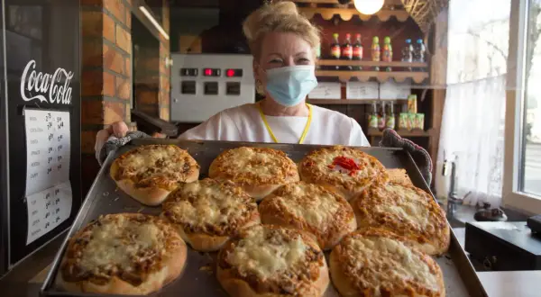 Uśmiechnięta sprzedawczyni w maseczce prezentuje tacę z zapiekanymi pizzami. To może być najstarsza pizzeria w Polsce, gdzie tradycja spotyka się z nowoczesnością.